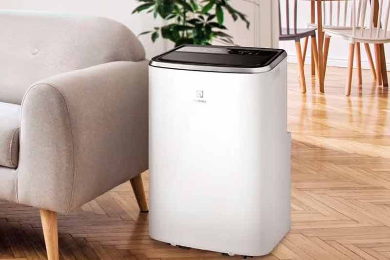 Portable air conditioner placed in a living room with chairs and a sofa behind it.