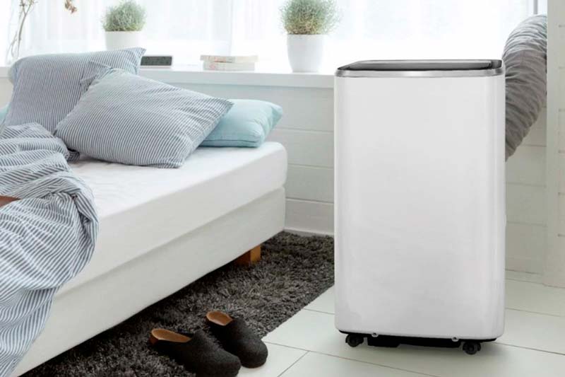Portable air conditioner installed next to a bed with slippers on the floor on a carpet.