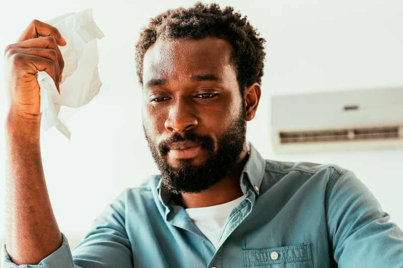 Man Sweating in apartment with air conditioner in the wall behind him
